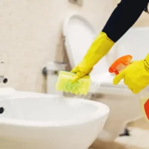 Cleaner with yellow gloves and a sponge spraying a bathroom